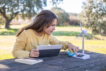 Girl engaging with a small wind turbine model and using a digital tablet on a wooden table, studying environmental science and sustainable energy future while enjoying nature