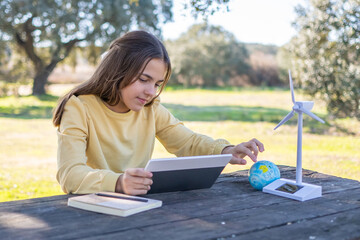 Fototapeta premium Young girl engaging in outdoor education, learning about eco-friendly concepts like renewable energy and sustainability using a tablet, a mini globe, and a wind turbine model