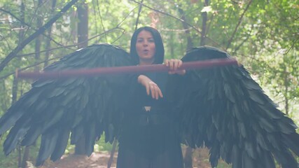 Young woman with black wings rotates staff gracefully in green forest, sunlight glowing from behind, expression full of allure and power, blending mystery, fantasy, and natural elegance