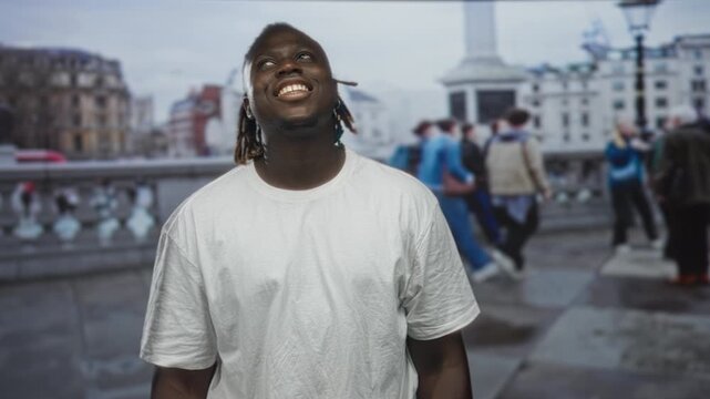 Man looking up and smiling while standing on street near bridge and monument in urban square wearing white t shirt with dreadlocks; joy.
