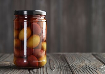 A glass jar filled with colorful pickled plums standing on a rustic wooden table. The preserved fruit is visible through the clear liquid, with red and yellow tones creating a natural, organic look.