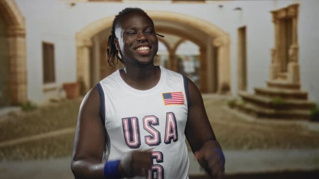 Man points index fingers upward on a cobbled street in an old town, smiling and wearing a usa basketball jersey with flag patch and blue wristbands; joy.