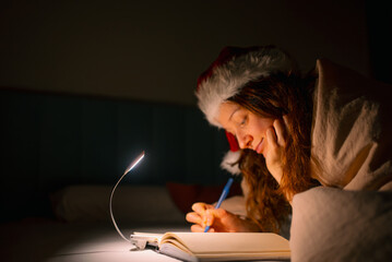Woman wearing a warm santa hat and blanket writing in a notebook by the light of a book lamp,...