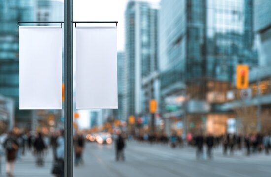 Two clean blank white banners hang from city streetlight pole. Noisy urban street forms backdrop with blurred modern glass buildings, many people walking. Perfect empty advertising space for product