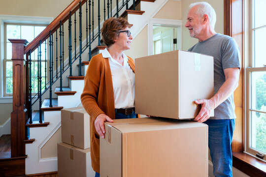 Caucasian senior man and Caucasian senior woman carrying cardboard boxes together near staircase, smiling and making eye contact, moving into new home, teamwork and relocation concept