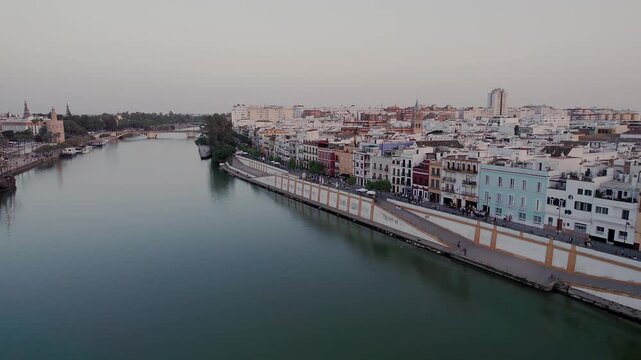 Guadalquivir River separating Triana district on left from Paseo de Crist&oacute;bal Col&oacute;n embankment on right, with Torre del Oro and Puente de Triana in background of Seville cityscape, low aerial, Spain