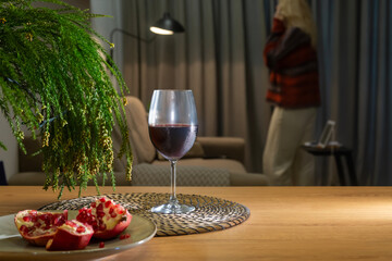 A glass of red wine, pomegranate and green fir branches on a wooden table against a blurred background of a room with a talking on the phone woman