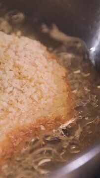 Close-up of breaded pork cutlet frying in hot oil in a stainless steel pan in a home kitchen, vertical format