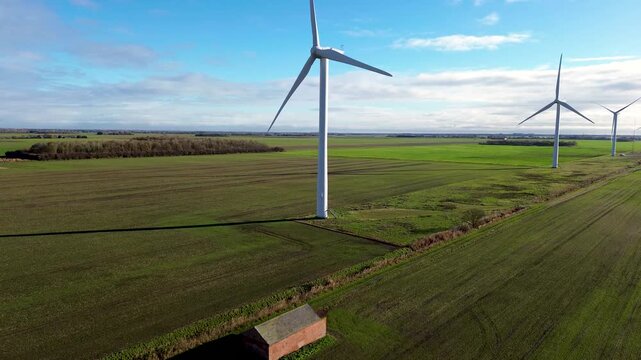 Close-up drone shot of a single wind turbine towering over expansive green farmland with a small farm building under a partly cloudy sky