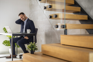 Business professional experiencing discomfort while working at desk near modern staircase in bright office setting