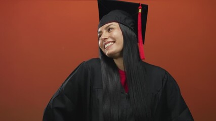 Young hispanic woman smiling in graduation uniform with cap in front of orange background, celebrating her achievement joyfully.
