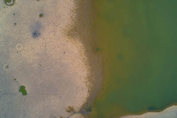 Top-down drone shot capturing the texture of dry cracked earth meeting murky green water. The receding shoreline and small toxic puddles illustrate the severity of the climate crisis and drought.