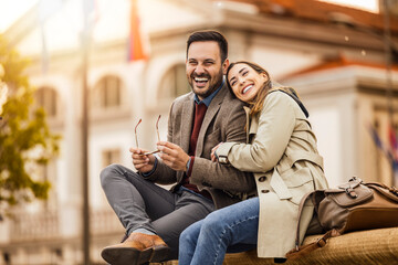 Couple enjoying a sunny afternoon together while sitting on a bench in a city square surrounded by...