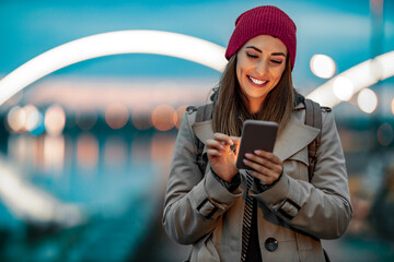 Smiling woman using smartphone by the river at sunset in a cozy winter outfit