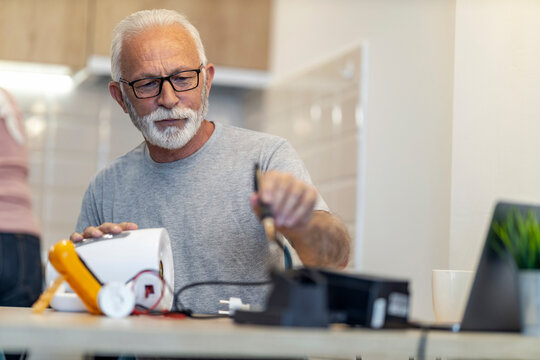 Senior man repairs electronic device at home while sitting at dining table in bright kitchen