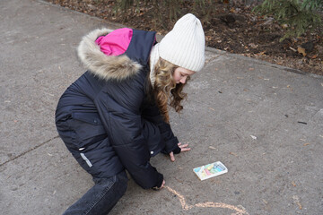 Creative young girl in a warm winter coat drawing with colorful chalk on a sidewalk.