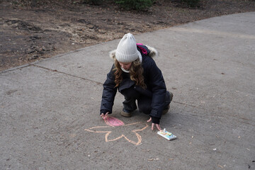 Fototapeta premium Child in a warm hat and jacket drawing a chalk flower on an asphalt pavement outdoors.