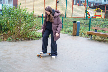 Young woman in a warm jacket learning to balance on a skateboard on a wet city sidewalk.