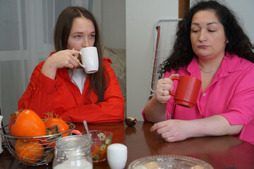 A young woman and a middle-aged woman sit together at a table drinking from mugs with pensive expressions.