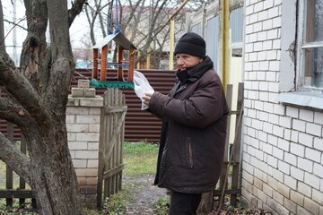 Elderly man feeding birds in his backyard, putting seeds into a handmade feeder.