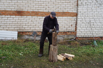 Man in warm clothes splitting firewood with an axe in a rural backyard.