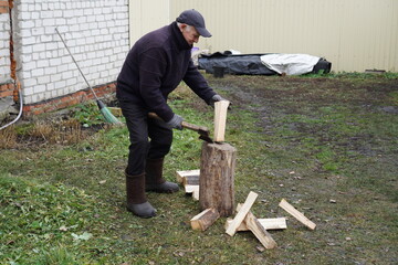 Active senior man in a cap and boots chopping firewood with an axe in the backyard on a cool day.