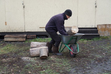 Man loading large log into a wheelbarrow, preparing firewood for winter in a rural backyard.