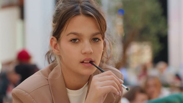 caucasian teen girl vaping at cafe, pensive closeup portrait on busy outdoor terrace with soft bokeh, natural daylight and casual jacket, expressive facial gestures, handheld vape device, onlookers