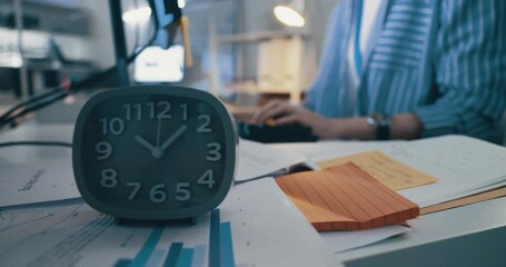 Close-up of a desk clock showing late night hours with a blurred worker typing in the background.
