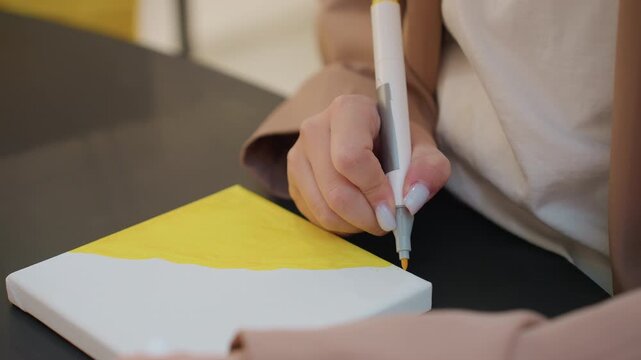 sketching vibrant canvas with precision, closeup of hand refining artwork on yellow surface, focused artist using marker to perfect details on colorful art project in serene studio environment