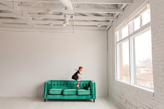 Young girl mid-jump on teal couch near large bright windows