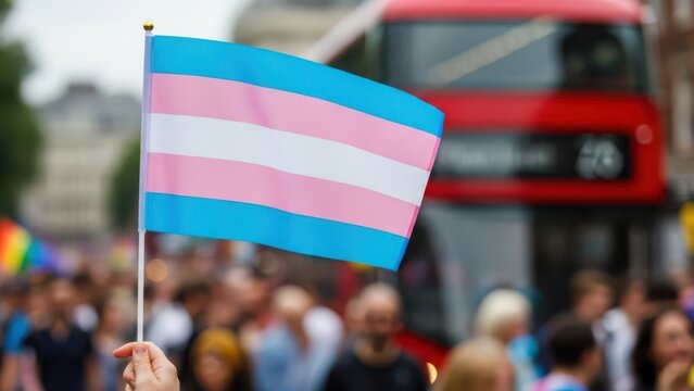 Transgender pride flag waved at a rally with a blurred red bus and crowd in the background