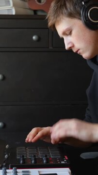 Young caucasian man wearing professional headphones concentrating on pressing pads of a Midi drum machine controller, producing music at home, beatmaking and audio engineering, vertical shot