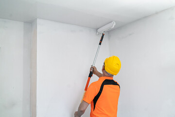 A construction worker paints a ceiling with a wide, specialized paint roller. Apartment renovation.