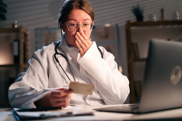 Confused doctor woman with laptop and credit card planning budget working during night shift in hospital. Financial problem concept