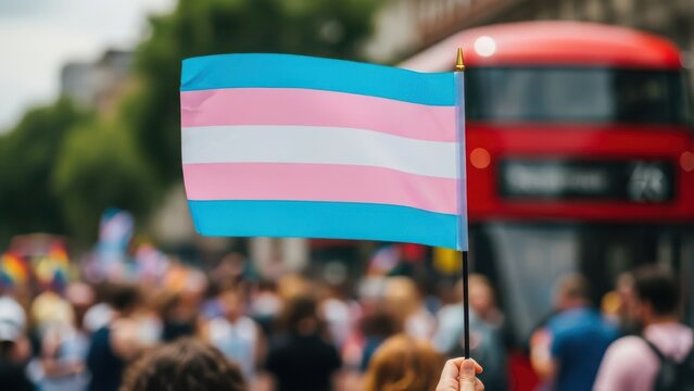 A transgender pride flag is held aloft at a pride event with a blurred crowd and red bus in the background