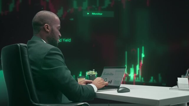 Focused Young Black Male Trader in Fintech Workspace Studies Live Candlestick Charts on His Laptop, Tracking  Stock, Managing Investments, Analysing Financial Data Wall. 360 Degree Tracking Arc Shot