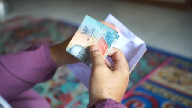 A woman's hand puts fifty thousand rupiah notes into a plain white envelope.