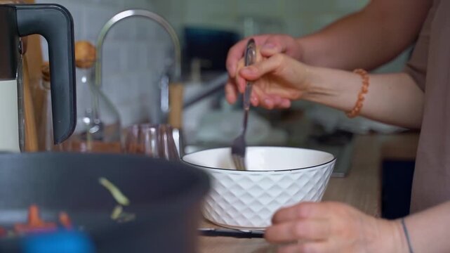 A woman's hand beats eggs in a bowl. Two hands, a woman's and a man's, beat eggs in a white deep plate.
