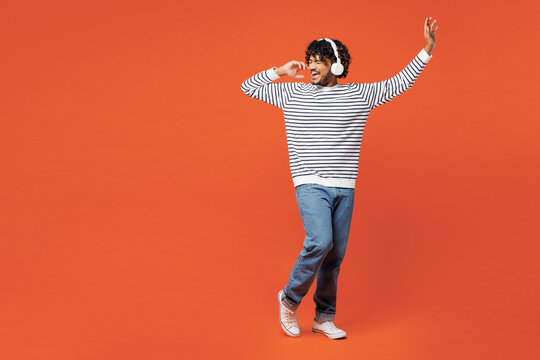 Full body young smiling happy Indian man he wear striped sweatshirt casual clothes listen to music in headphones raise up hands dance isolated on plain red orange background studio. Lifestyle concept. - Powered by Adobe