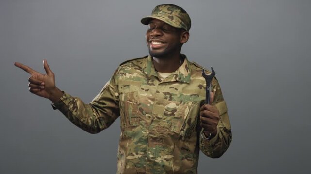 Soldier in camouflage uniform smiling, pointing finger with left hand and holding large wrench at chest in studio; pride duty.