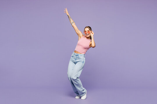Full body young happy cheerful fun cool woman she wear pink tank top casual clothes sunglasses dance raise up hands isolated on plain pastel light purple background studio portrait. Lifestyle concept.