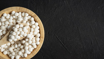 Organic tapioca pearls in wooden bamboo bowl on black table top view with copy space. Spoon scooping white dry tapioca pearls, cooking ingredient, gluten free food, healthy eating.