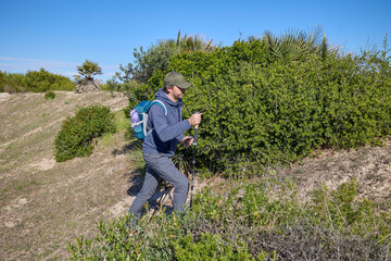 Man hiking uphill with trekking poles on a dry coastal trail surrounded by bushes on a sunny day. Outdoor activity, exercise and adventure in nature.