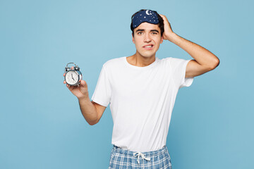 Calm young sad shocked man he wear pyjamas jam sleep eye mask rest relax at home hold in hand clock...