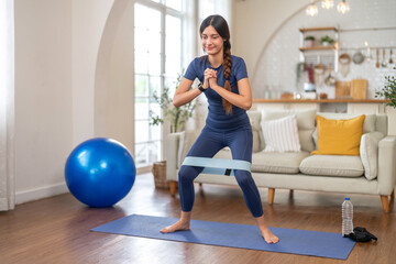 Fit smiling young woman doing exercise resistance band squats at home on yoga mat at home workout, healthy lifestyle, cardio fitness and strength training, bodyweight training and warm up, wellness