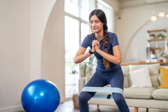 Fit smiling young woman doing exercise resistance band squats at home on yoga mat at home workout, healthy lifestyle, cardio fitness and strength training, bodyweight training and warm up, wellness - Powered by Adobe