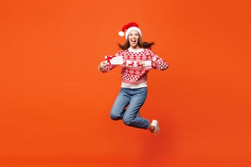 Obraz premium Full body merry young woman in red Christmas sweater Santa hat posing jump high hold gift coupon voucher card for store isolated on plain orange background Happy New Year celebration holiday concept