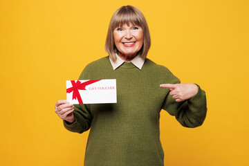 Elderly blonde woman wear green sweater casual clothes hold point finger on gift certificate coupon voucher card for store isolated on plain yellow orange background studio portrait Lifestyle concept