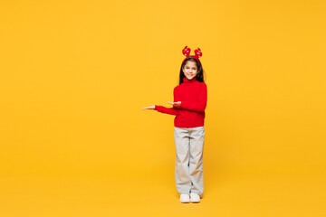 Full body merry little girl wearing red clothes fun decorative deer horns on head posing point...